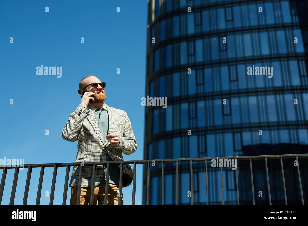 Positivo bello mirato barbuto manager in occhiali da sole in piedi contro il grattacielo e avente la conversazione telefonica Foto Stock