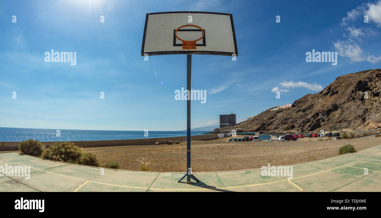 La pallacanestro tavola spinale con cestello sul vecchio campo sportivo. Un enorme edificio abbandonato di fronte all'oceano nel lontano sullo sfondo. Tenerife. Grandangolo, f Foto Stock