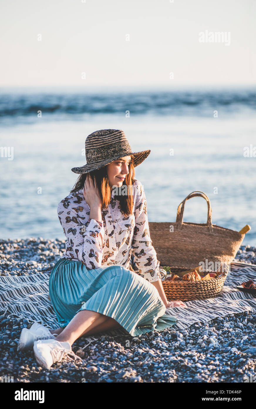 Estate picnic sulla spiaggia al tramonto. Giovane donna seduta su una coperta avente fine settimana picnic all'aperto al mare con frutta fresca di stagione e il vassoio del gustoso app Foto Stock