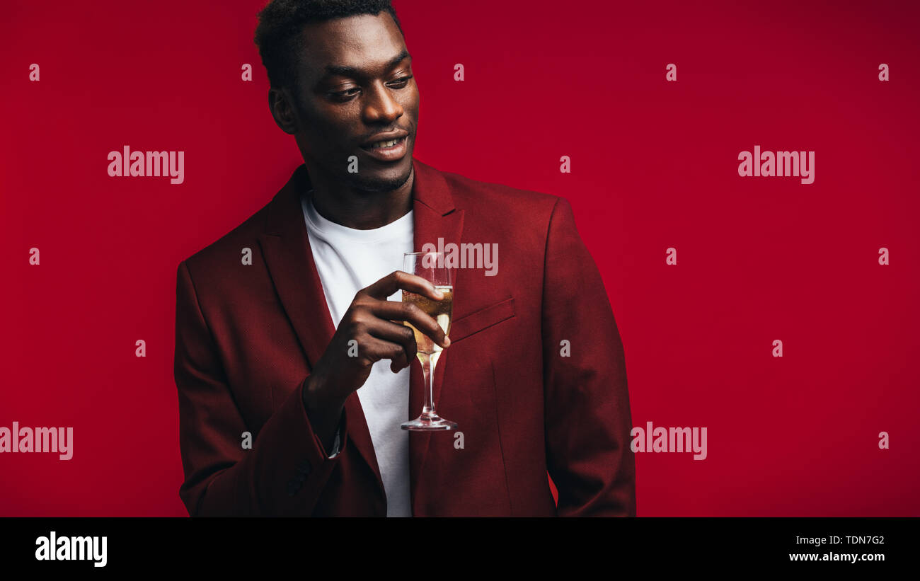 Elegante uomo africano con un bicchiere di champagne che guarda lontano a copiare lo spazio. Uomo in camicia rossa avente lo champagne contro sfondo colorato. Foto Stock