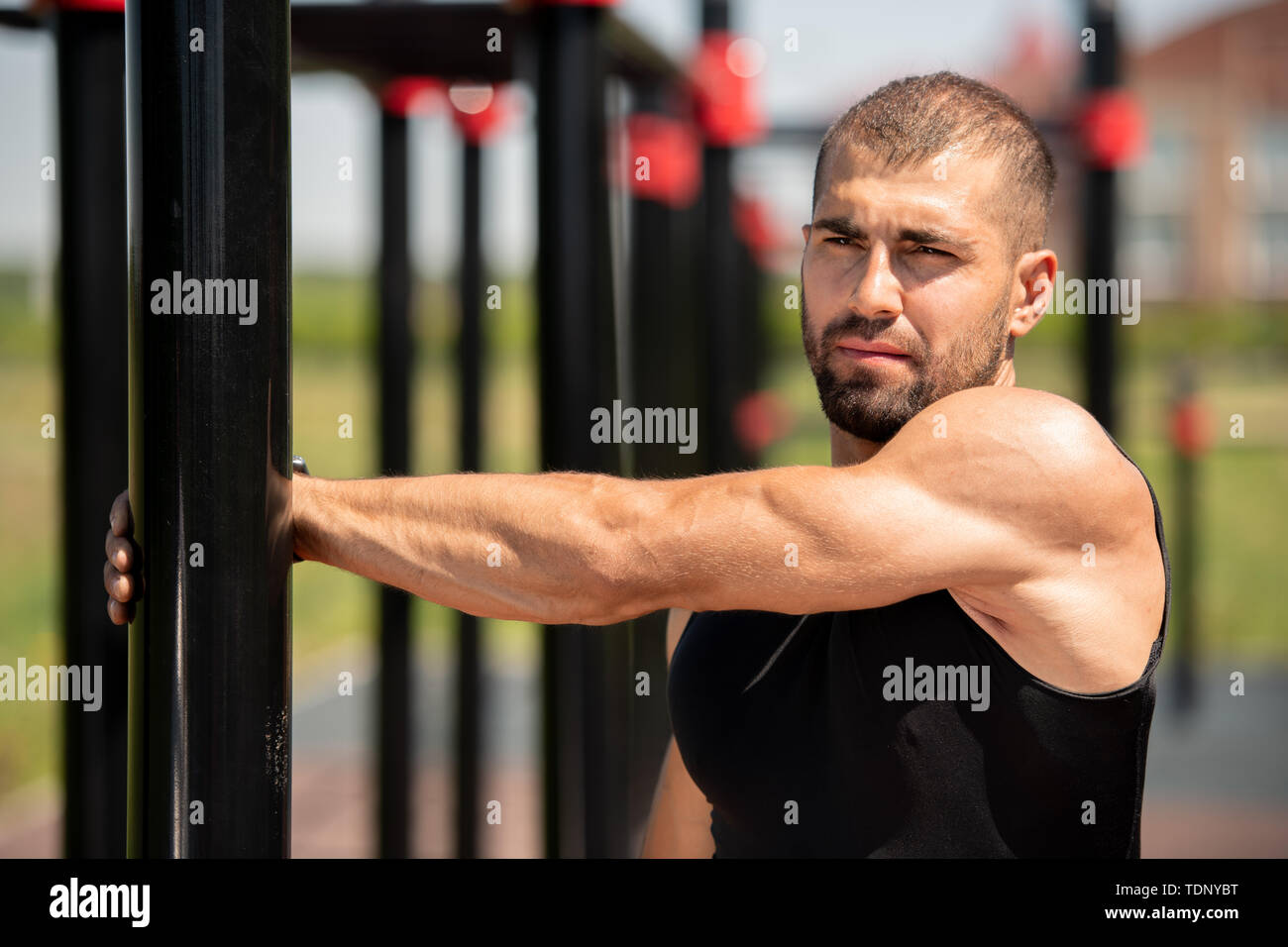 Guy muscolare nel gilet nero azienda dal bar sport mentre si estende il braccio durante gli allenamenti su strutture sportive all'aperto Foto Stock