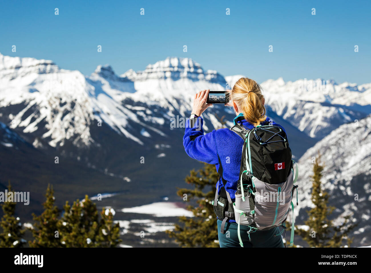 Escursionista femmina sulla cima della montagna con un telefono cellulare di scattare una foto della coperta di neve la gamma della montagna con il blu del cielo; Banff, Alberta, Canada Foto Stock