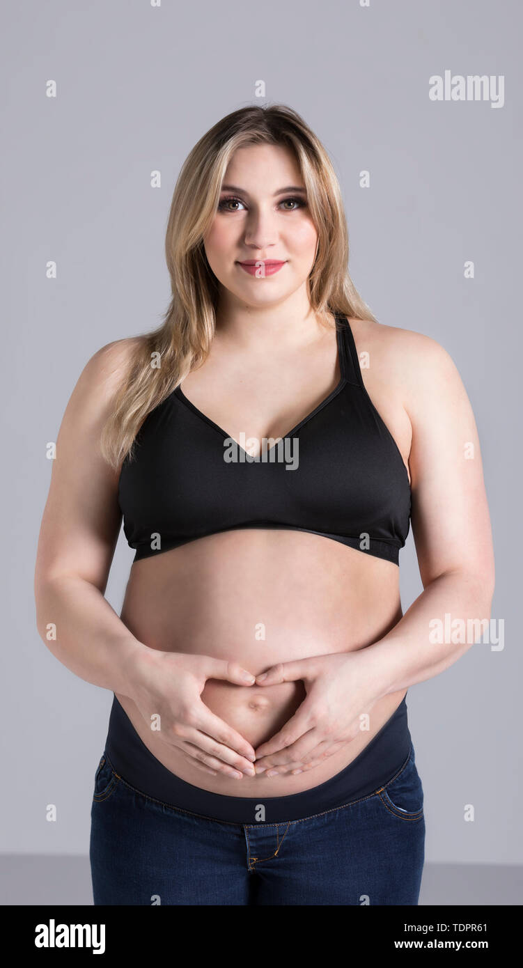 Una giovane donna incinta mentre tiene il suo ventre in studio e in posa per la fotocamera mentre si effettua una forma di cuore oltre il suo bambino Foto Stock