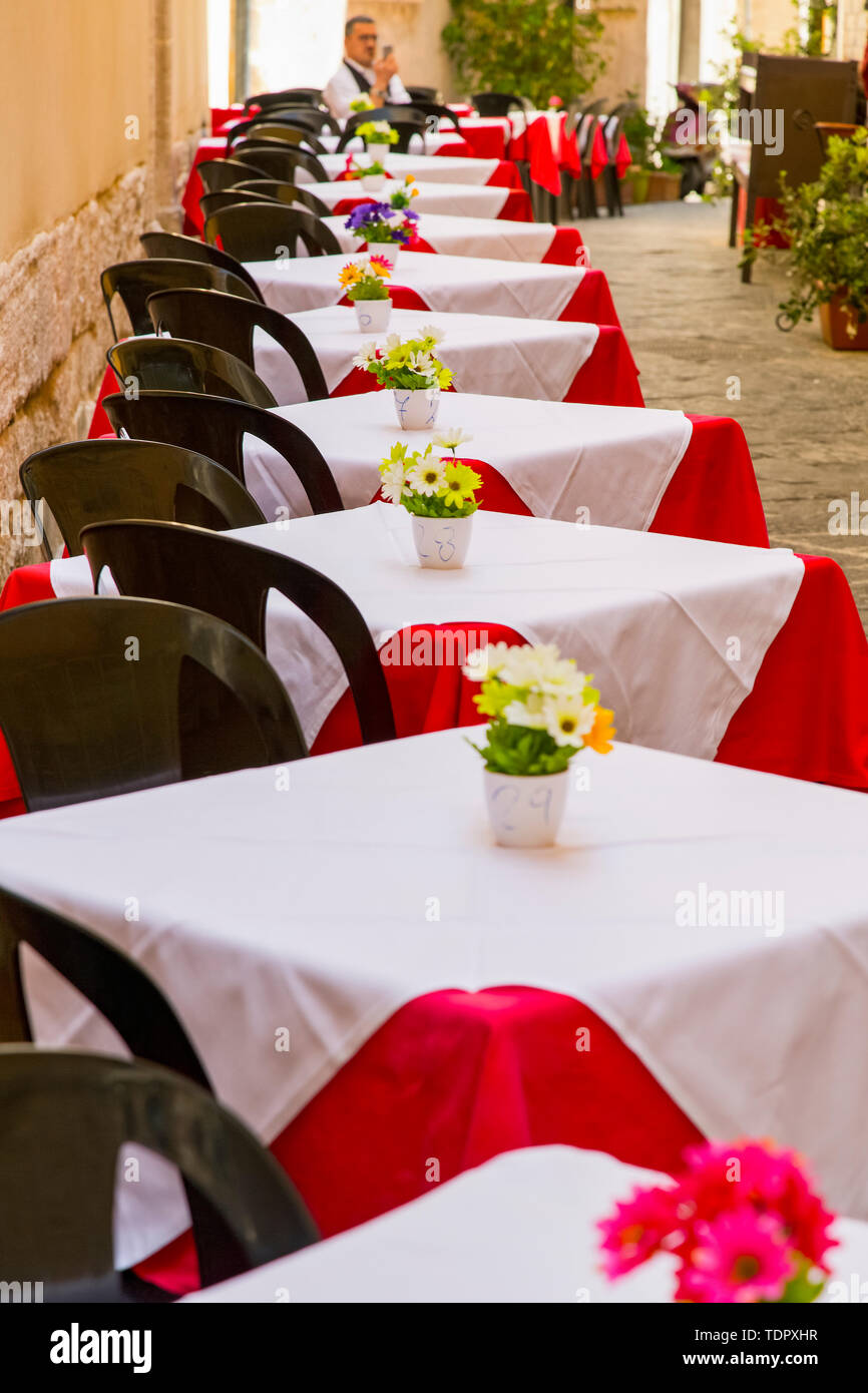 Set di tabelle su un patio esterno in un ristorante in Italia con un maschio di soggiorno turistico e utilizzando il suo smart phone; Siracusa, Sicilia, Ortigia, Italia Foto Stock