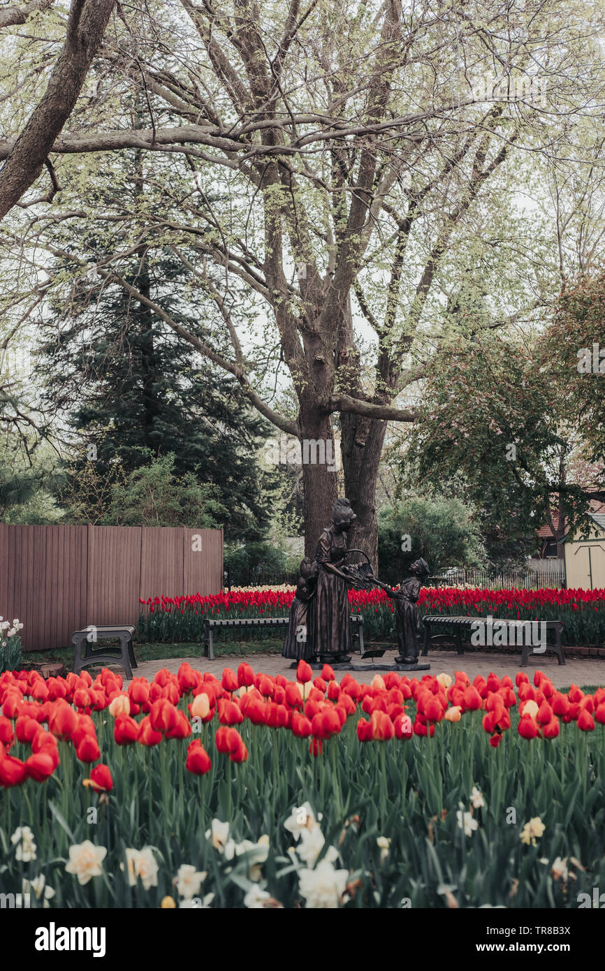 Pella, Iowa, Stati Uniti d'America - 2 Maggio 2019: scultura di una donna Olandese e due bambini con un cesto di tulipani nei giardini di Scholte. Letti di rosso e tulipani gialli. Foto Stock