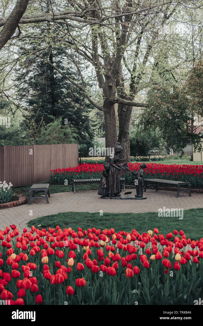 Pella, Iowa, Stati Uniti d'America - 2 Maggio 2019: scultura di una donna Olandese e due bambini con un cesto di tulipani nei giardini di Scholte. Letti di rosso e tulipani gialli. Foto Stock