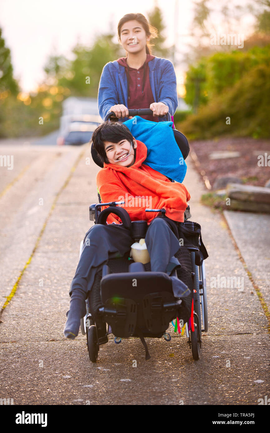 Big Sister spingendo disabilitato piccolo fratello in carrozzella intorno al quartiere, ridendo e sorridente Foto Stock