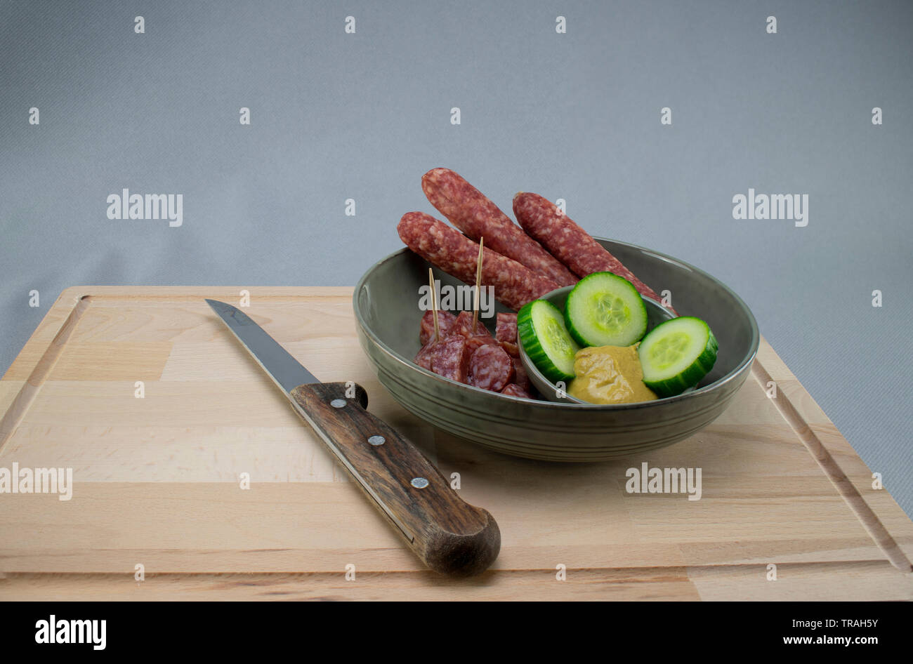 Una piccola ciotola con carne di maiale le fette di salsiccia di sottaceti Senape e su una tavola di legno con un coltello affilato e uno sfondo grigio Foto Stock