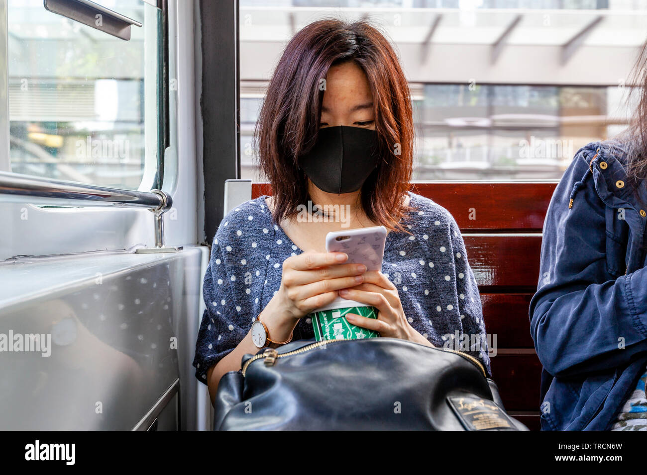 Una donna che indossa una maschera facciale seduto su un tram guardando il suo smartphone, Hong Kong, Cina Foto Stock