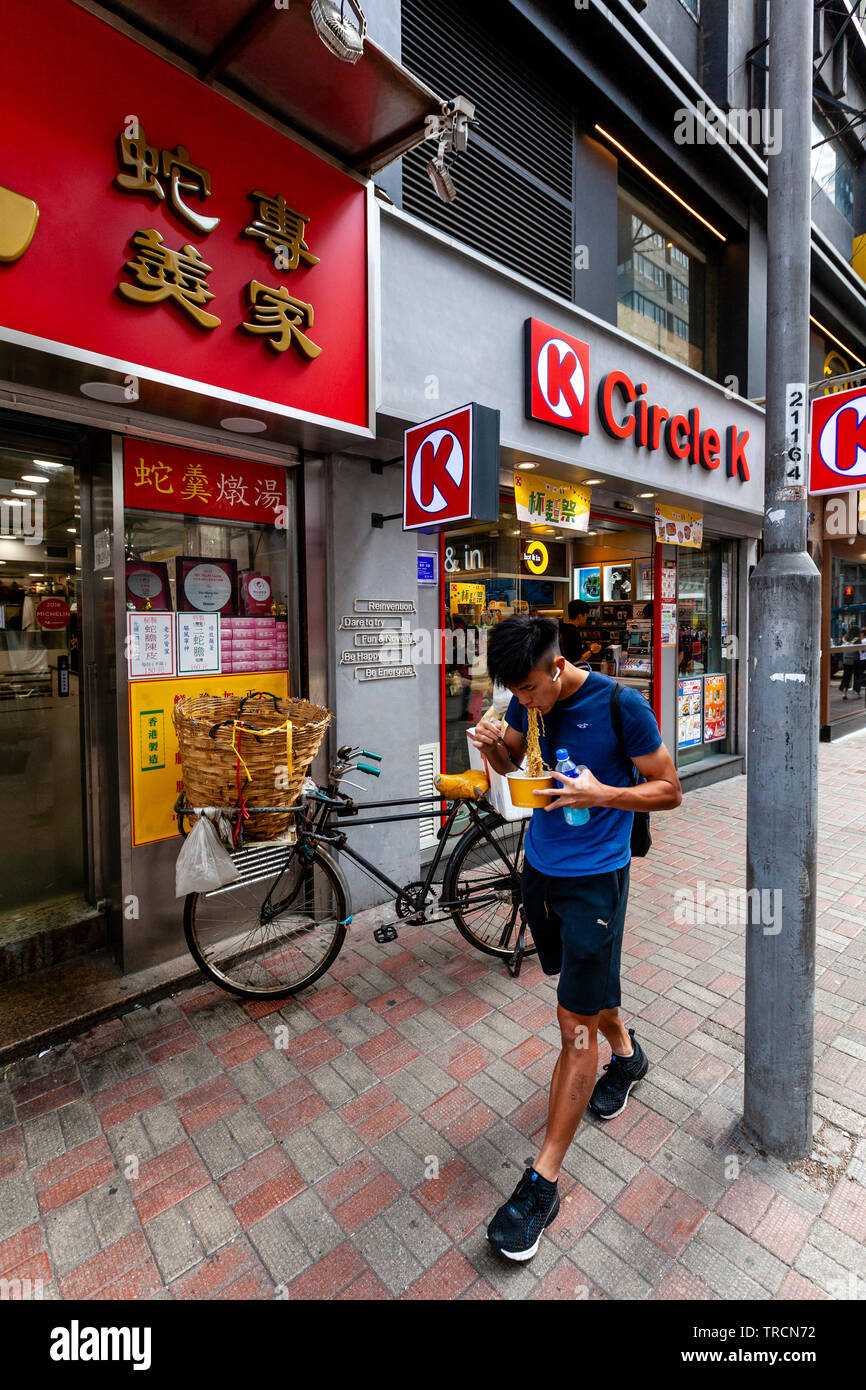 Un giovane uomo cammina per la strada mangiare tagliatelle, Hong Kong, Cina Foto Stock