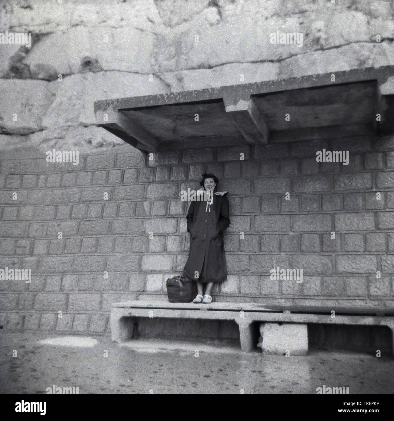 1950s, storica, al mare, una donna che indossa un cappotto in piedi su una panca, contro una parete di scogliera sotto il tetto di un rifugio in cemento, Inghilterra, Regno Unito. Foto Stock