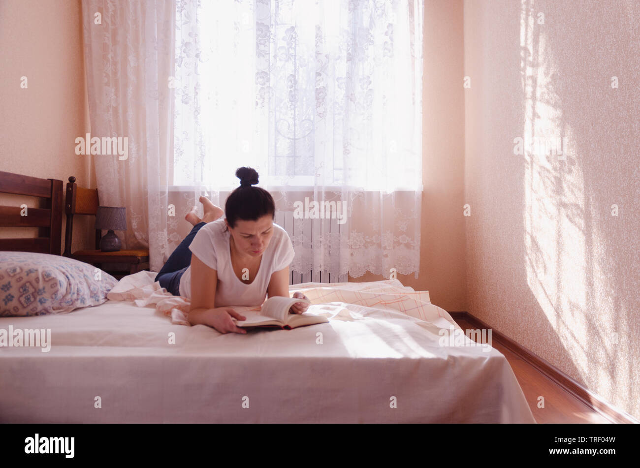 Un felice giovane donna leggendo un libro in una accogliente camera da letto nella mattina di sole. Foto Stock