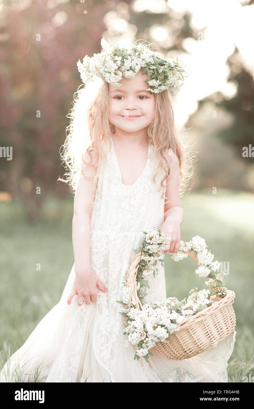 Carino kid girl 4-5 anno vecchio con cesto di fiori indossando ghirlanda floreale all'esterno. Guardando alla fotocamera. Infanzia. Giorno di nozze. Foto Stock