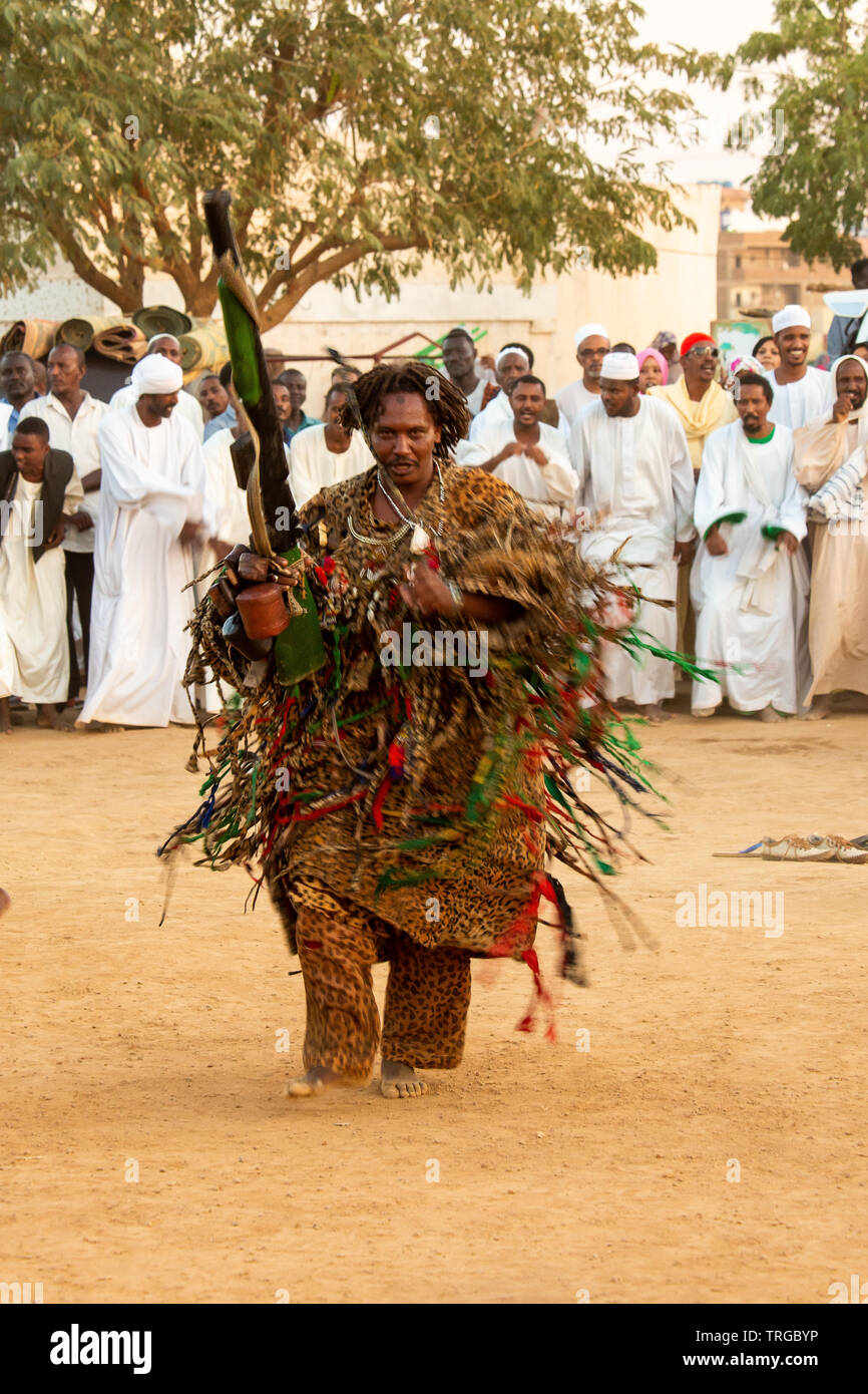 Sufi dreadlocked con capelli in pesanti costume bordati con imitazione legno fucile a derviscio armato cerimonia prima del tramonto a Omdurman, Sudan Foto Stock