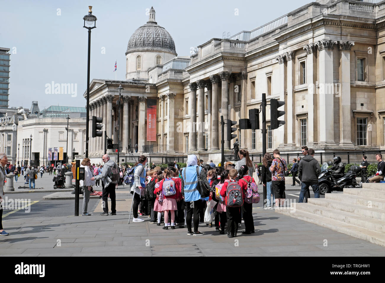 La gente che camminava in strada e un gruppo di studenti della scuola primaria gli alunni a visitare la Galleria Nazionale di Trafalgar Square a Londra England Regno Unito KATHY DEWITT Foto Stock