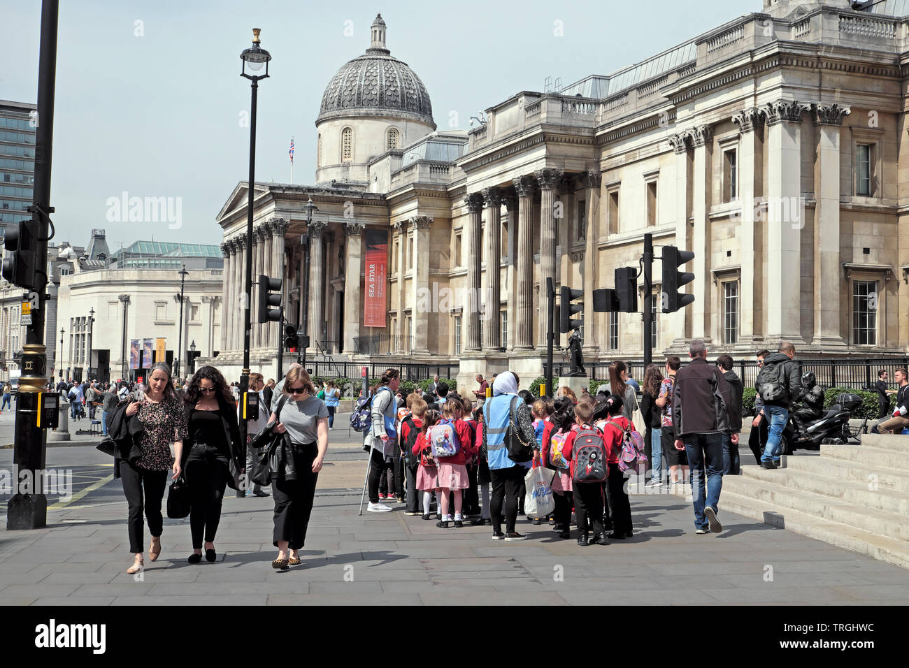 La gente che camminava in strada e un gruppo di studenti della scuola primaria gli alunni a visitare la Galleria Nazionale di Trafalgar Square a Londra England Regno Unito KATHY DEWITT Foto Stock