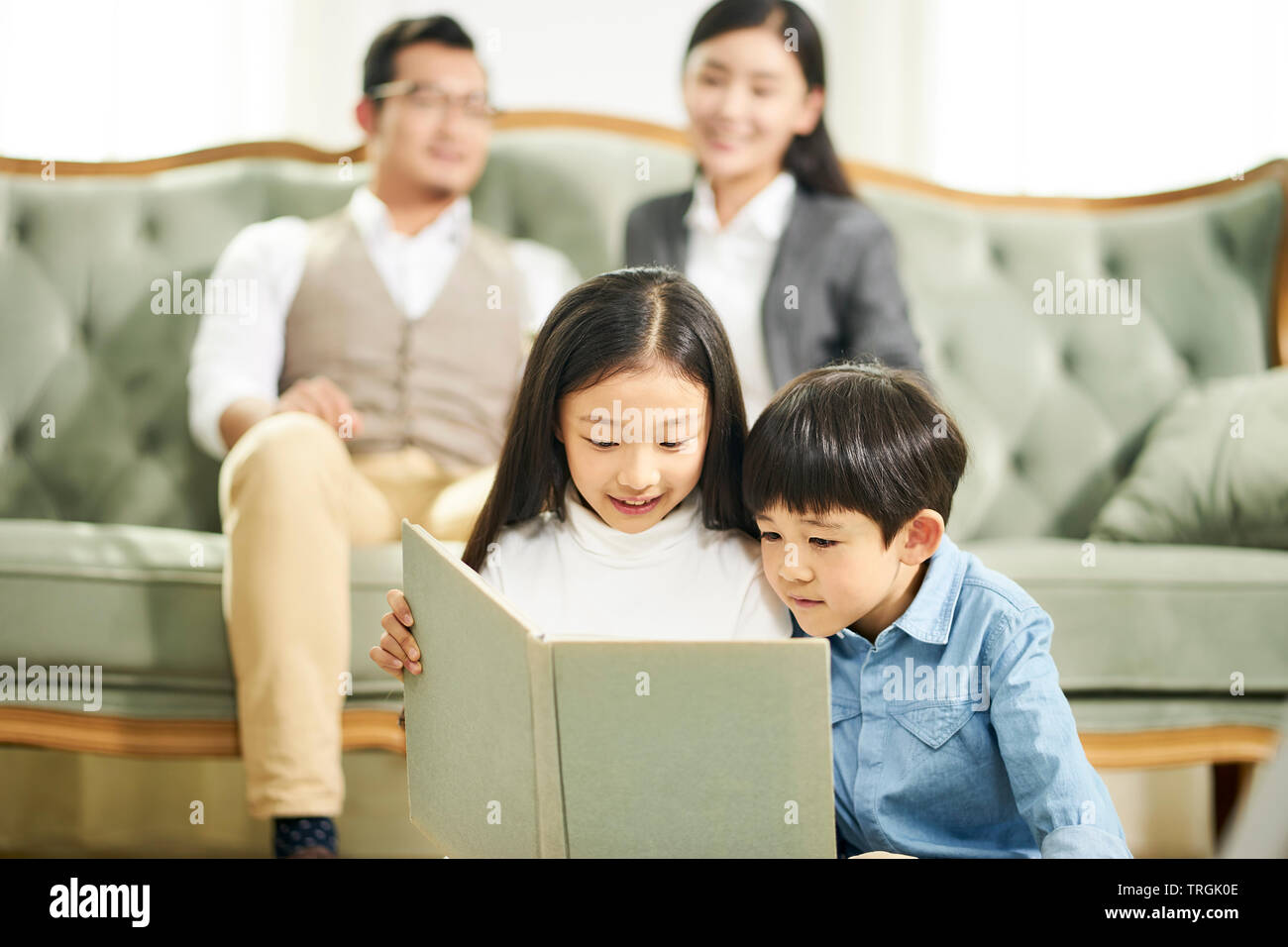 Due ragazzi asiatici del fratello e sorella seduta sul tappeto la lettura libro insieme a vivere in famiglia in camera con i genitori seduti sul divano in background. Foto Stock