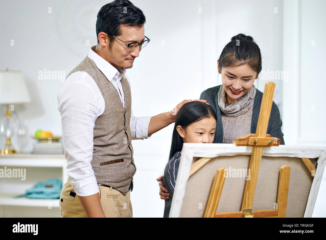 Bella ragazza asiatica con lunghi capelli neri facendo un dipinto su tela mentre i genitori di stare dietro a guardare. Foto Stock