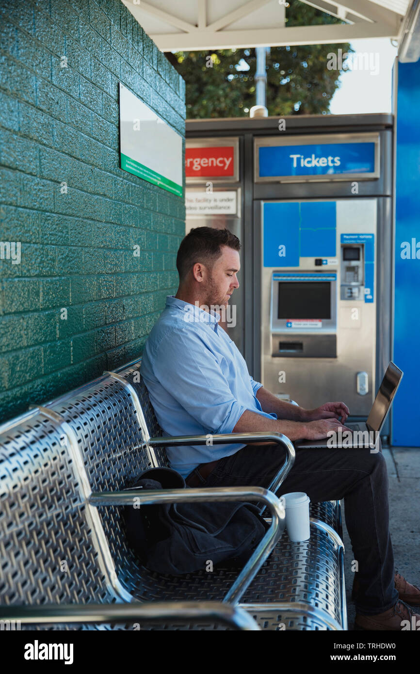Una vista laterale colpo di una metà degli adulti imprenditore caucasico lavorando sul suo computer portatile mentre si è seduti in attesa del treno alla stazione ferroviaria e piattaforma. Foto Stock
