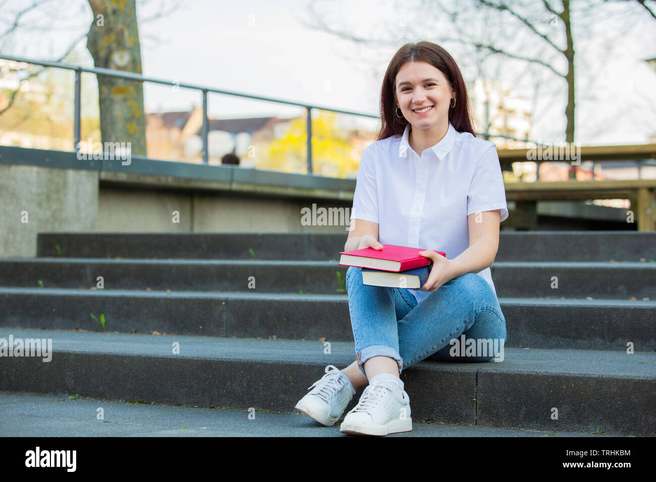Sorridente ritratto femminile di studente universitario seduti sulle scale del Campus con libri cercando di fotocamera. Studente fiducioso e sorridente holding boo Foto Stock