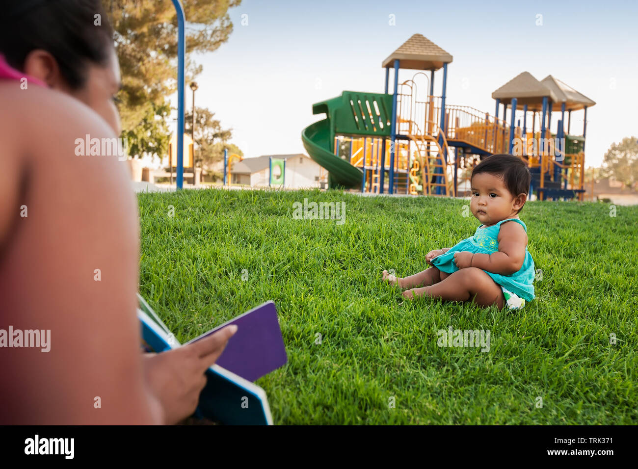 Una donna in possesso di un libro per bambini tenta di innestare l'attenzione di un bambino ragazza seduta nel prato di un parco giochi per bambini. Foto Stock