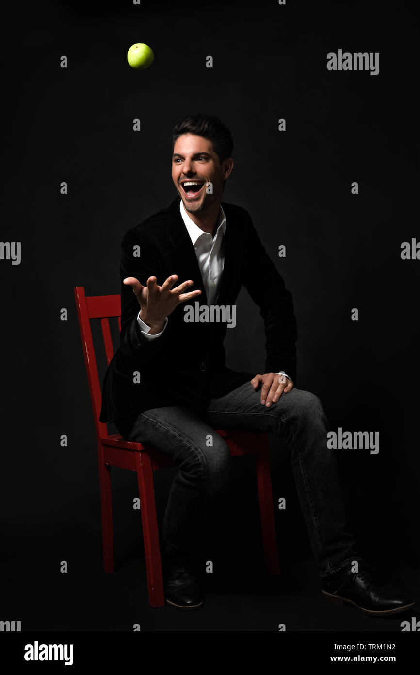 Una buona ricerca bianco modello maschio in un nero giacca formale, seduti su una sedia rossa, gettando una mela verde in aria, sorridente e la sensazione di essere eccitati! Foto Stock