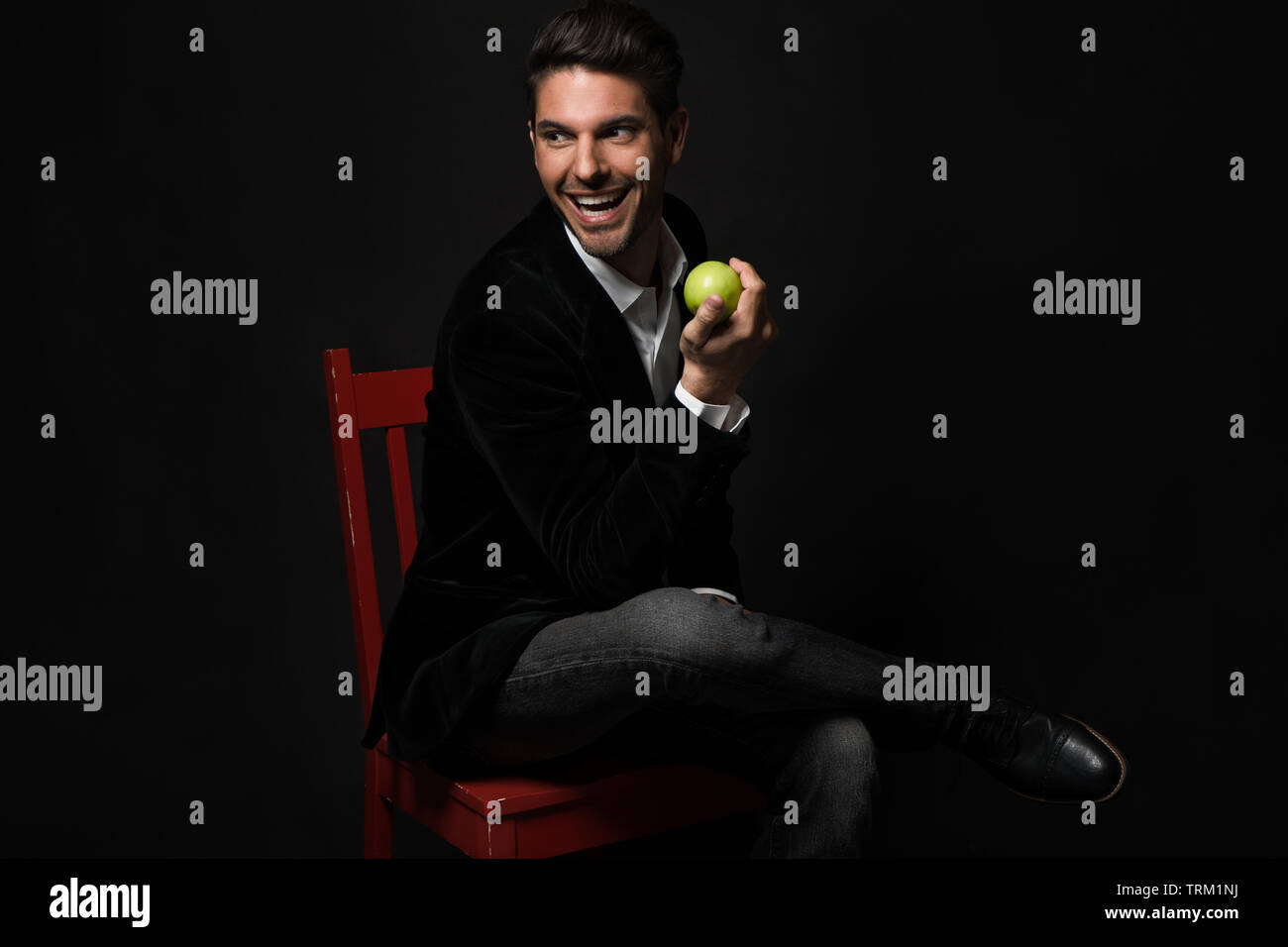 Una buona ricerca bianco modello maschio si siede con le gambe incrociate su una sedia rossa, tenendo una mela verde. Egli guarda lontano sorridente in un giocoso umore. Foto Stock