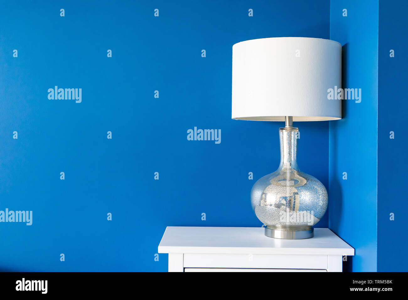 Vibrant Blue parete dipinta di bianco home decor accenti (un paralume bianco e argento metallico di supporto della lampada, su una notte bianca tabella). Spazio per testo e Foto Stock
