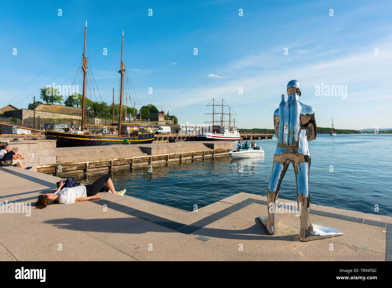 Oslo Norvegia estate, la vista di una donna a prendere il sole accanto a una scultura di un subacqueo in argento (Dykkar) su una banchina del porto (Aker Brygge) di Oslo. Foto Stock