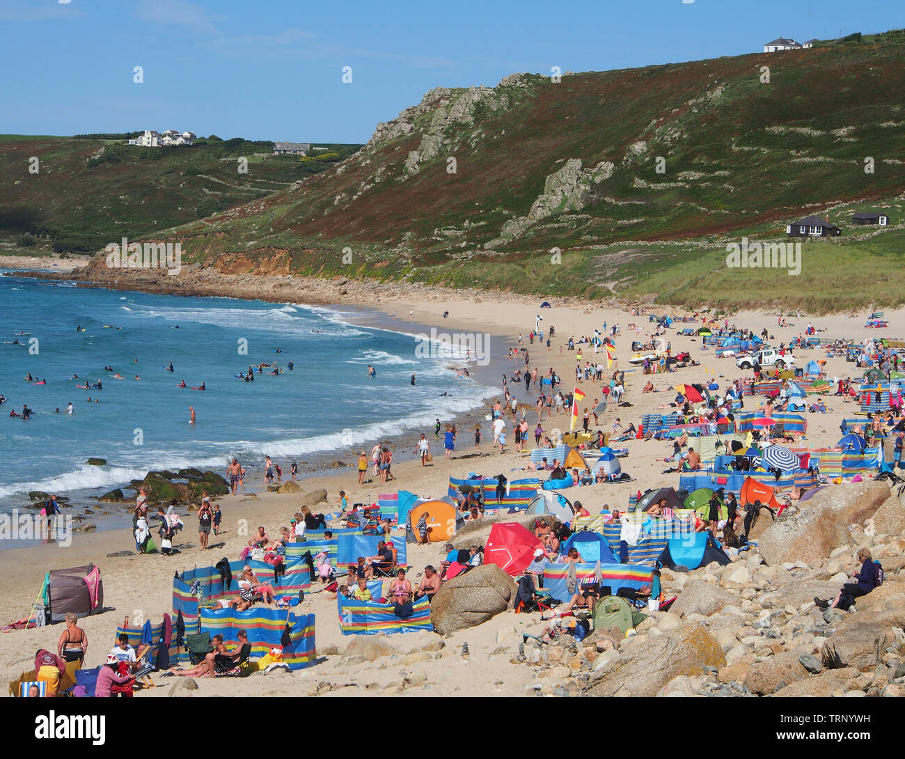 Folle di persone che si godono una giornata estiva soleggiata sulla spiaggia a Sennen Cove Cornwall Inghilterra, Regno Unito mostrando la spiaggia di sabbia e il mare blu sotto un cielo blu. Foto Stock