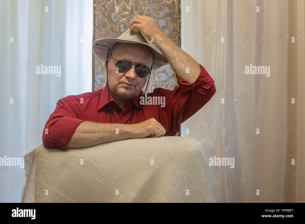 Ritratto di uomo maturo che indossa un cappello e occhiali da sole nero seduta e magra e il gomito sulla sedia con la schiena Foto Stock