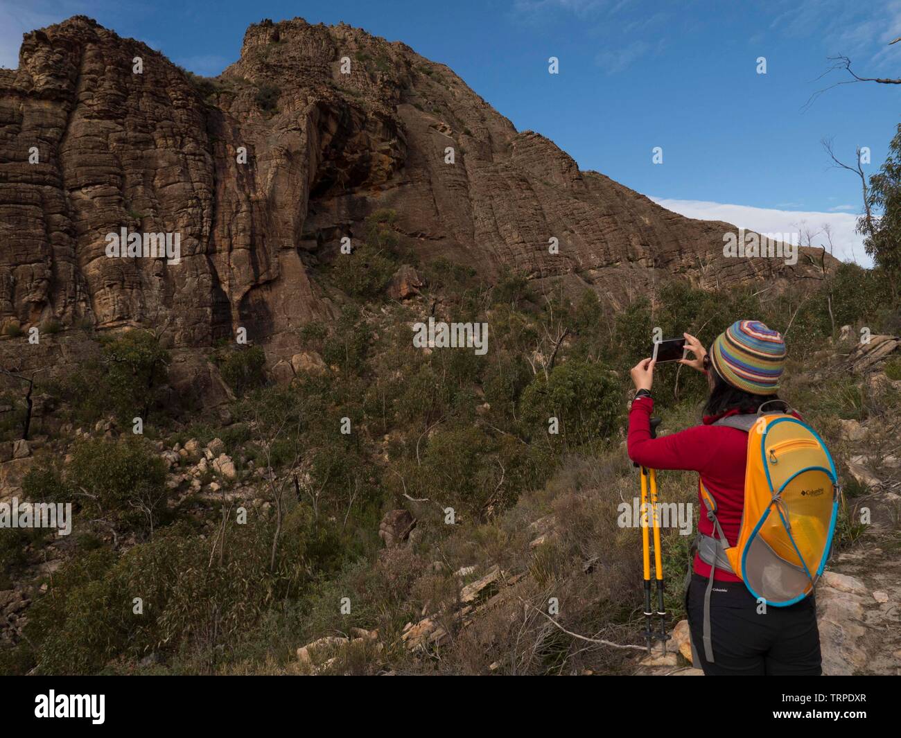 Giovane donna nella parte superiore rossa, pantalone nero e giallo day pack a scattare foto di formazioni rocciose nel Parco Nazionale di Grampians Australia con IPhone. Foto Stock