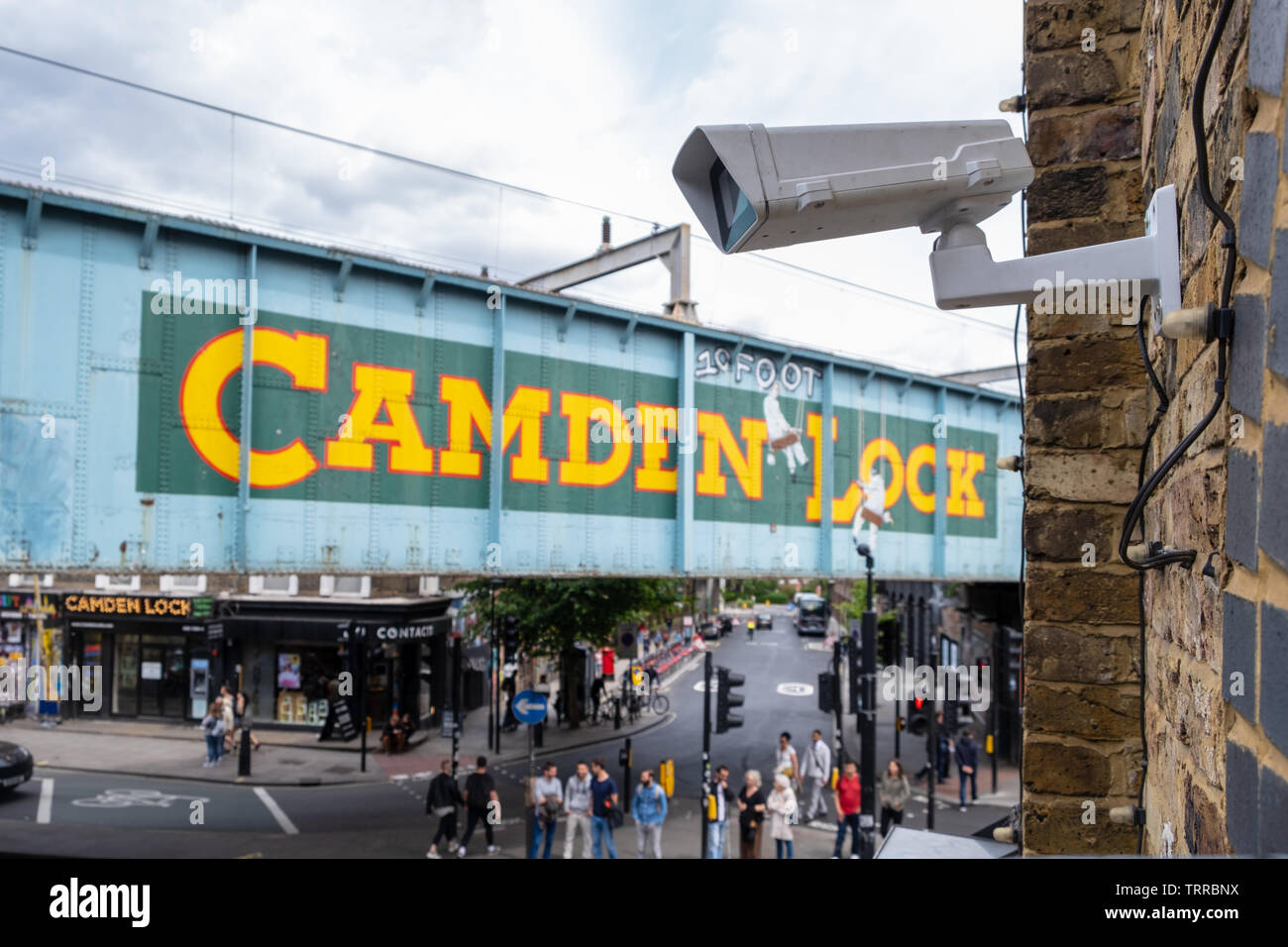 Sicurezza telecamera TVCC affacciato sul mercato di Camden e iconico dipinto di Camden Lock segno sul lato del ponte ferroviario a Londra con street e persone in Foto Stock