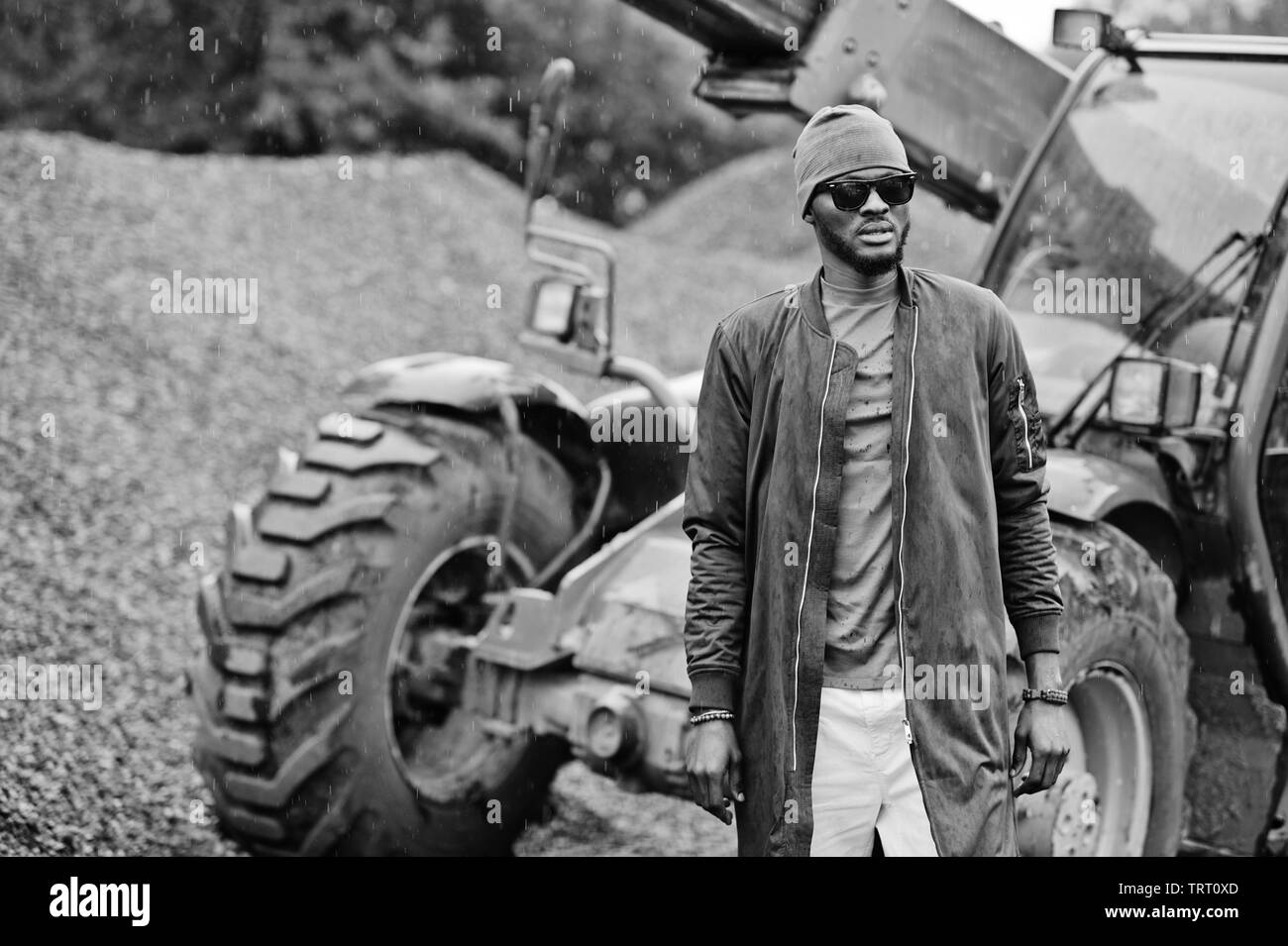 Elegante African American uomo nel cappello e occhiali da sole poste all'aperto sotto la pioggia contro il trattore con una benna. Foto Stock