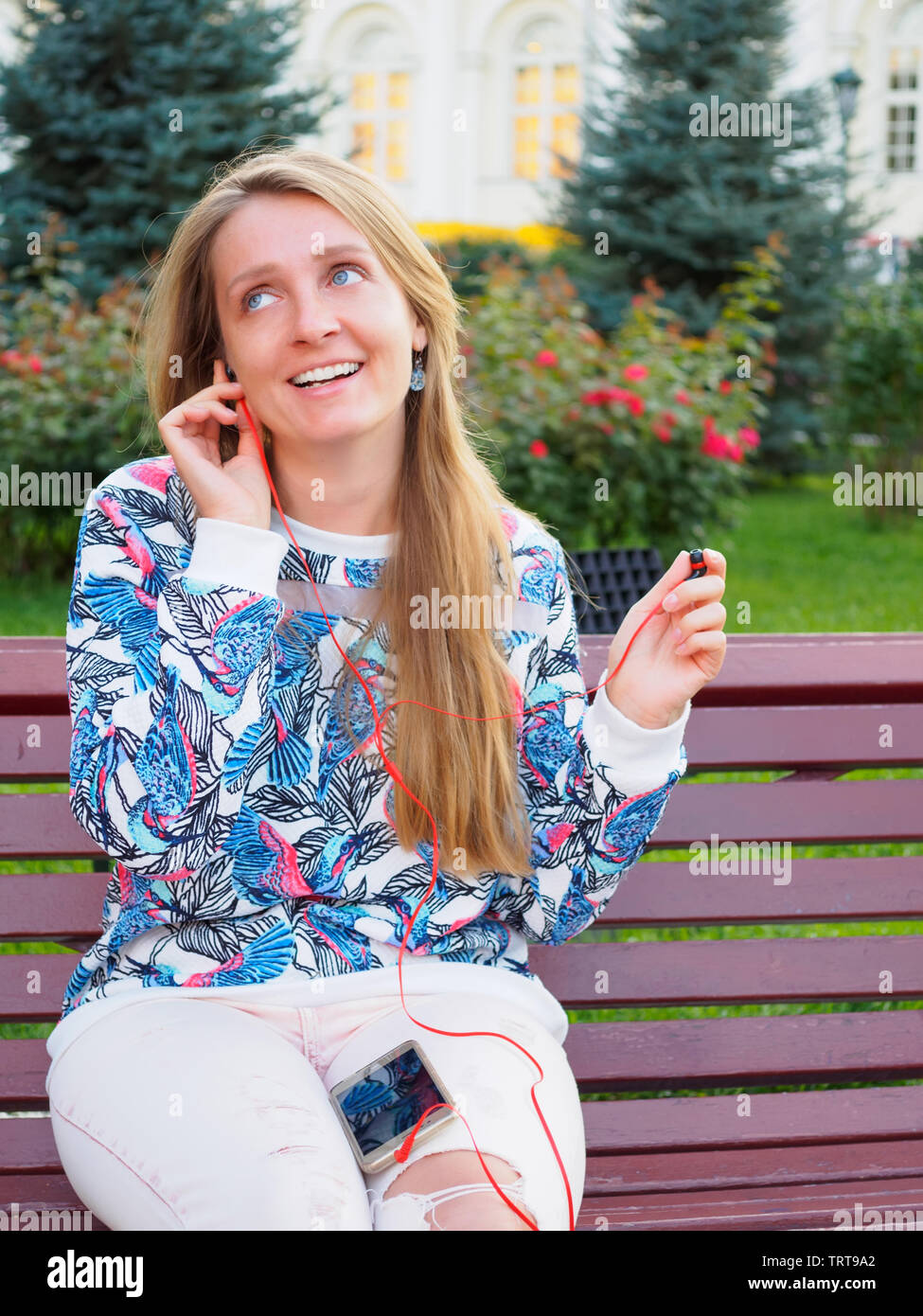 Giovane bella ragazza bionda seduta su una panchina nel parco con uno smartphone sorridente, parlando al telefono, ascolto di musica con un auricolare. Foto Stock