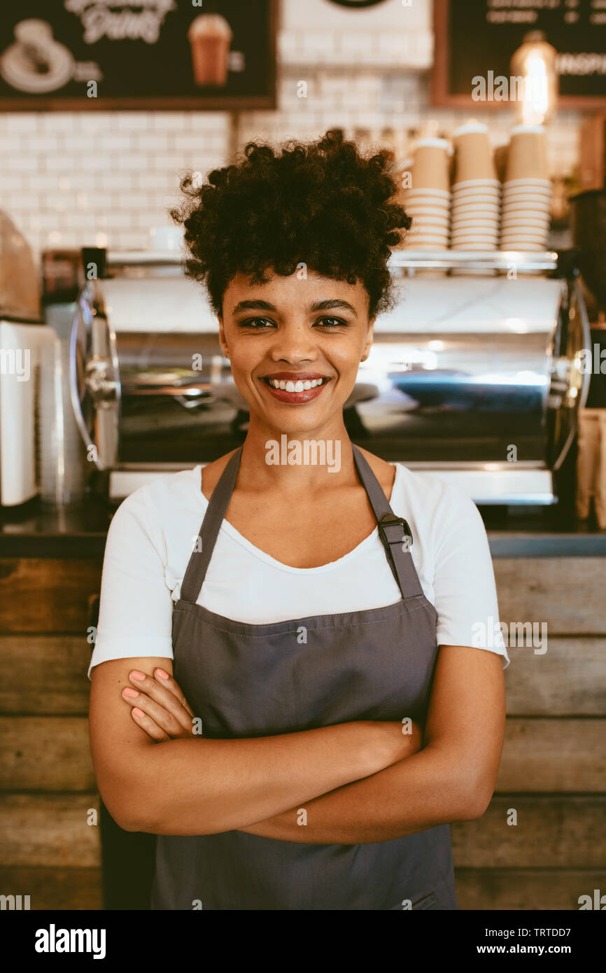 Fiducioso barista femmina in piedi di fronte ad un cafe contatore. Donna cafe proprietario nel grembiule guardando la fotocamera e sorridente con le braccia incrociate. Foto Stock