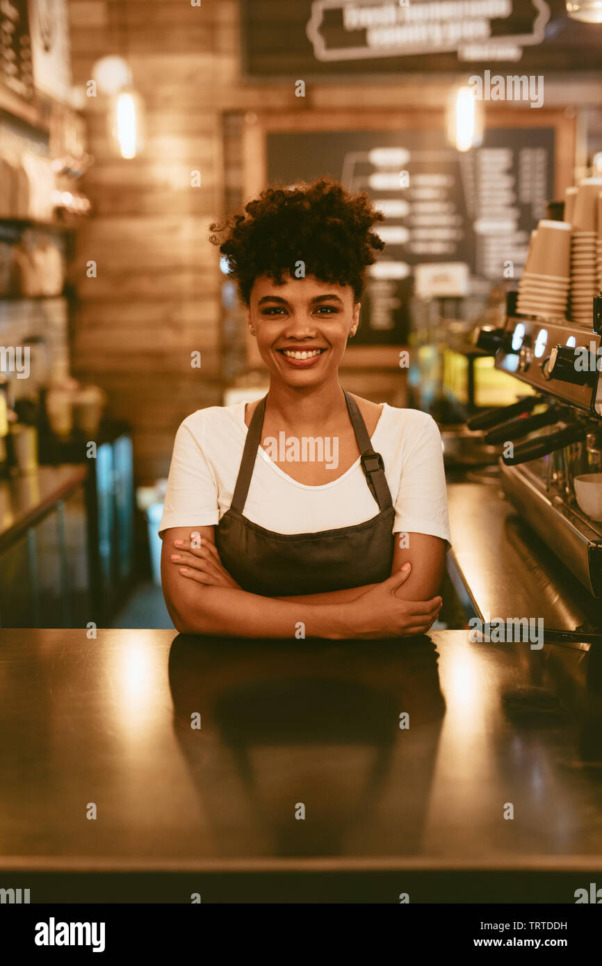 Sorridente donna africana nel grembiule permanente al cafe contatore. Fiducioso barista femmina in piedi dietro il contatore. Foto Stock
