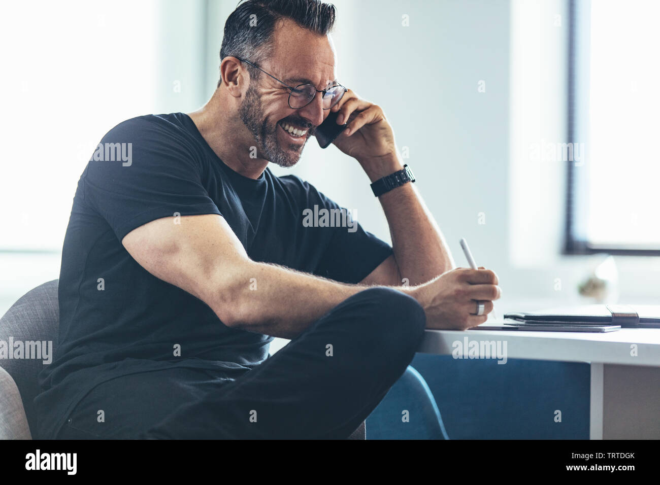 Imprenditore maturo parlando al cellulare e sorridente in ufficio. Imprenditore sorridente seduto alla sua scrivania e parlando sul suo telefono cellulare. Foto Stock