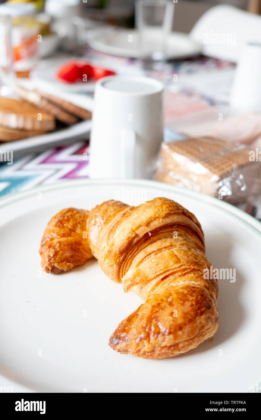 Primo piano di un croissant in una piastra su un tavolo per la colazione, accanto ad un bianco di ceramica pentola con il latte e alcuni vassoi con toast e affettati Foto Stock