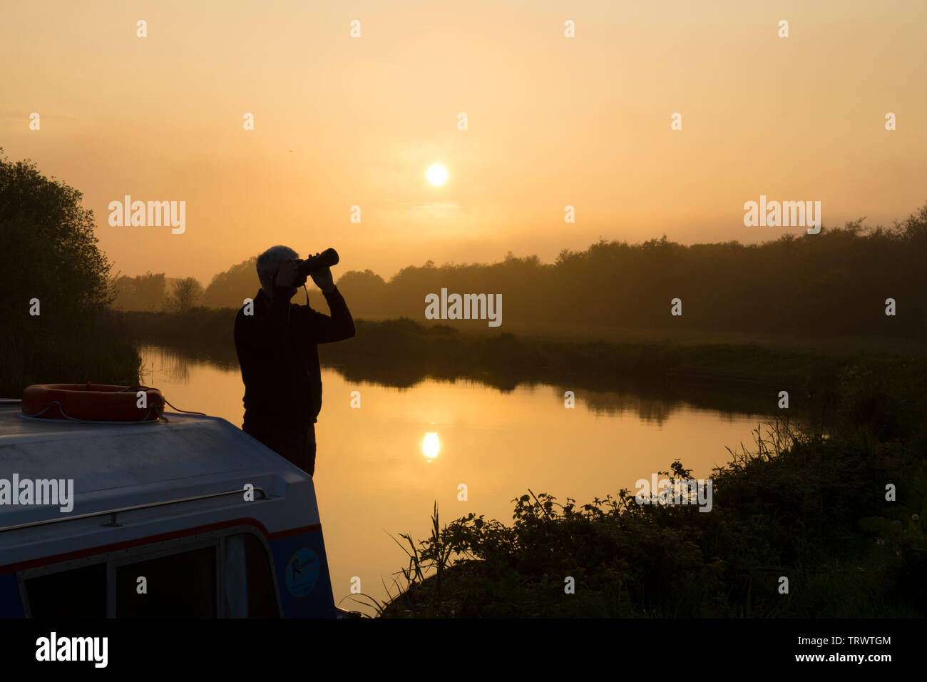 Uomo in vacanza sulla coperta della imbarcazione da diporto barca a fotografare un tramonto sul fiume Ant, il Parco Nazionale Broads del Norfolk, Regno Unito, maggio. Foto Stock
