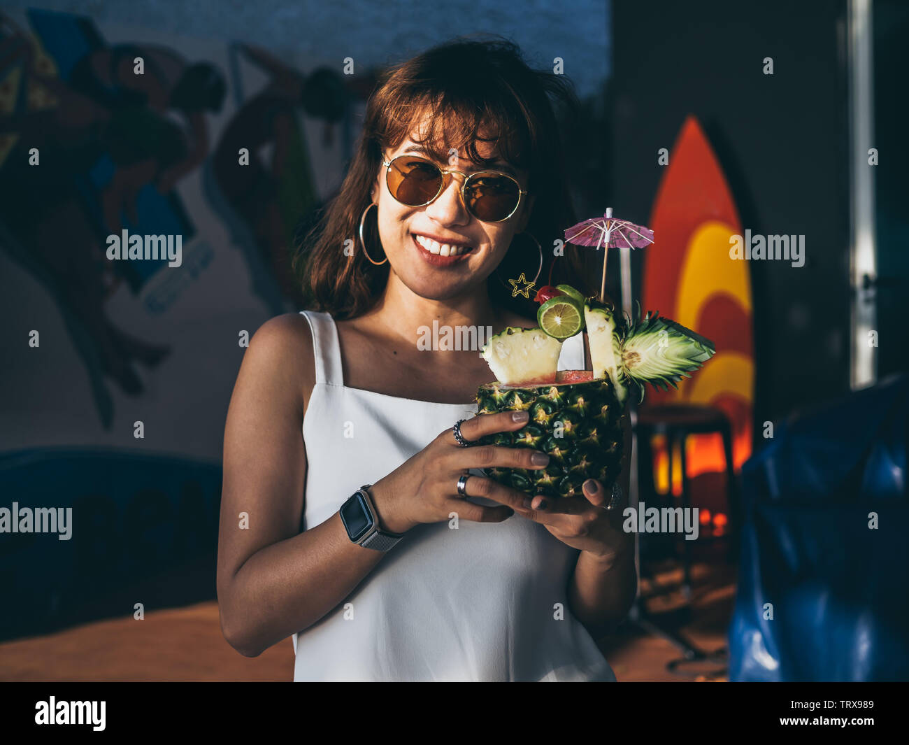 Felice bella donna asiatica che indossa senza maniche bianco e occhiali da sole tenendo fresco succo di ananas cocktail in estate. Foto Stock