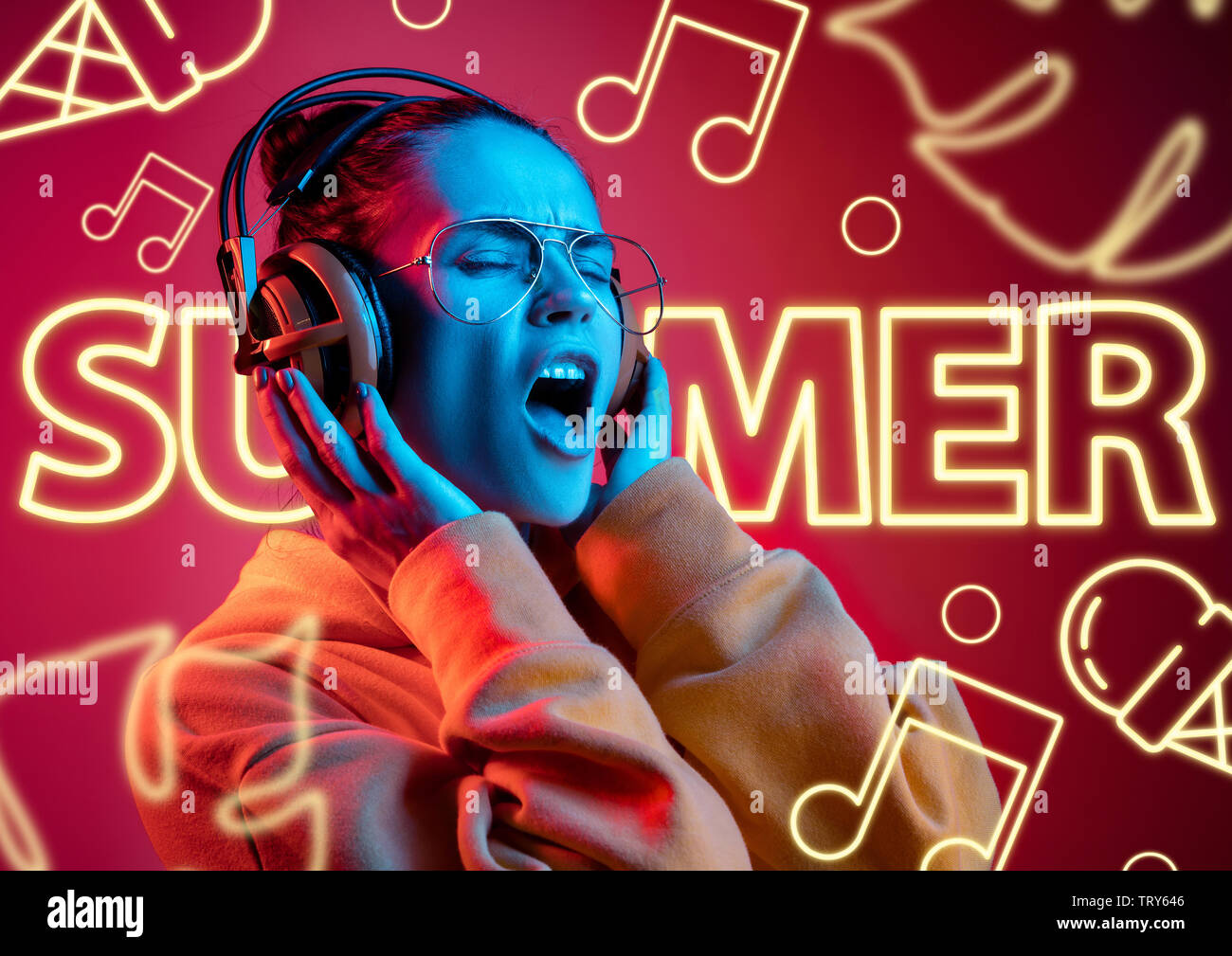 Modello femminile nel quartiere alla moda di luce al neon su sfondo rosso. Il design moderno. Bella giovane donna ascoltare musica con le cuffie e il canto. Concetto di espressione del viso, estate, vacanze, musica open-air. Foto Stock