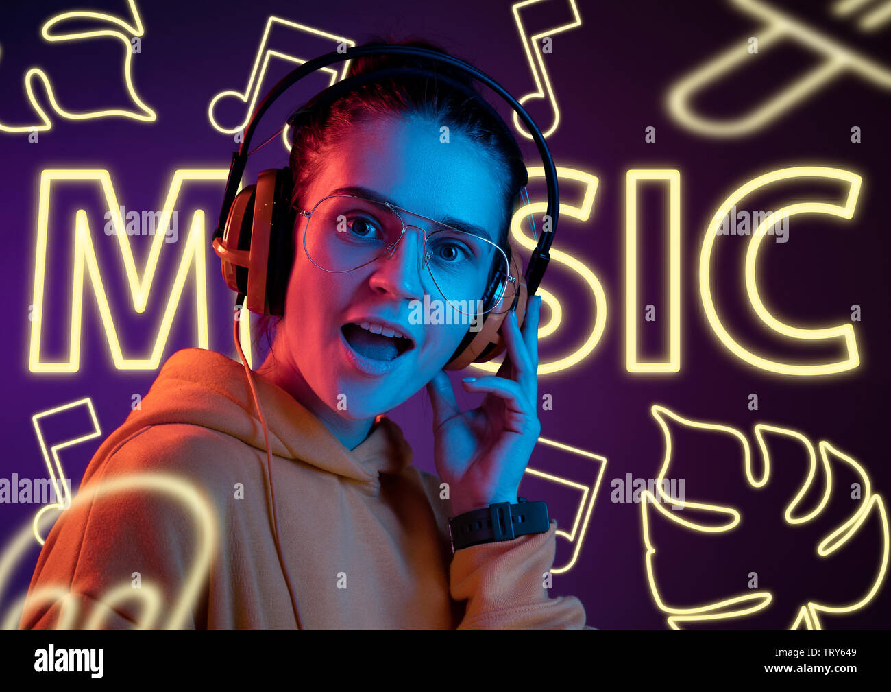 Modello femminile nel quartiere alla moda di luce al neon su sfondo viola. Il design moderno. Bella giovane donna ascoltare musica con le cuffie e il canto. Concetto di espressione del viso, estate, vacanze, musica open-air. Foto Stock