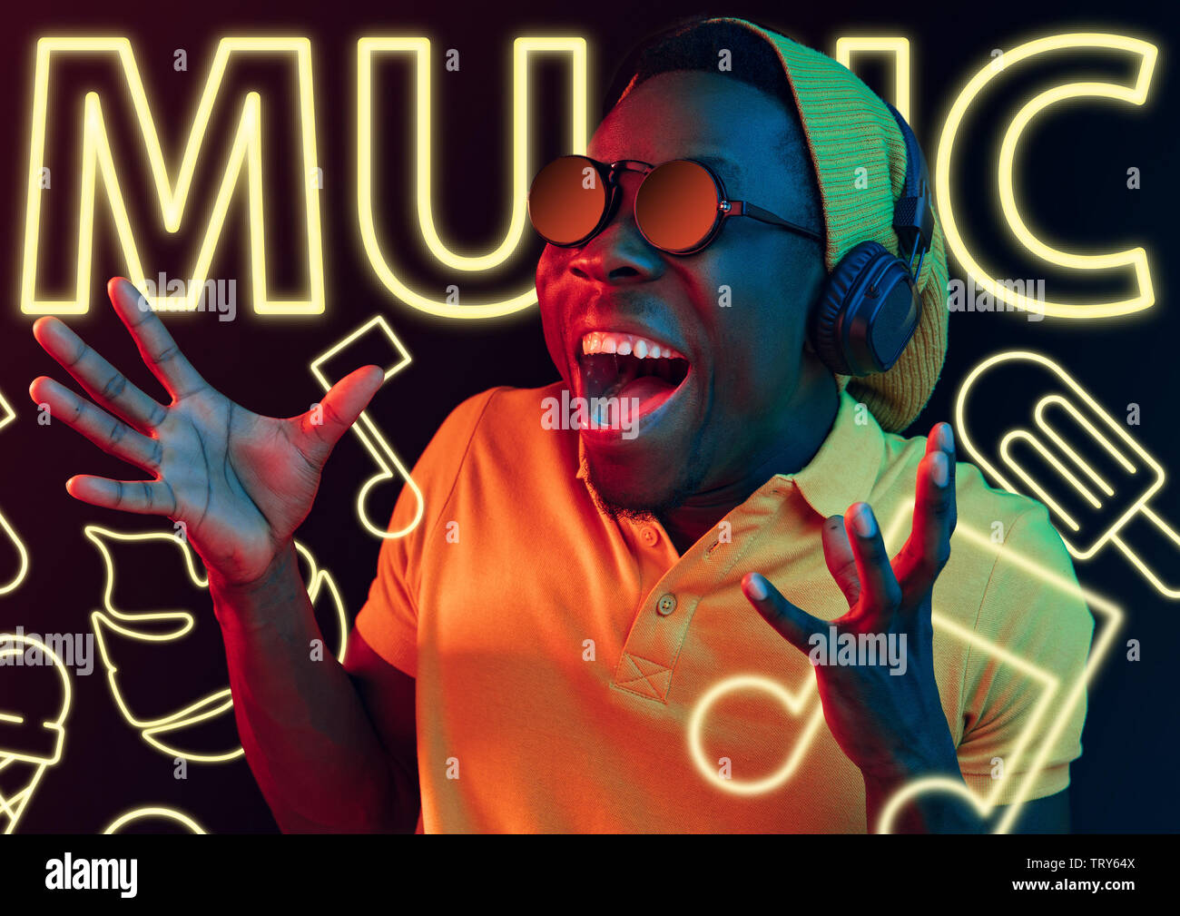 Bella giovane uomo su sfondo darkstudio in luce al neon. Il design moderno. Modello maschile in occhiali da sole con le cuffie sguardi stupiti. Concetto di espressione del viso, estate, vacanze, musica open-air. Foto Stock