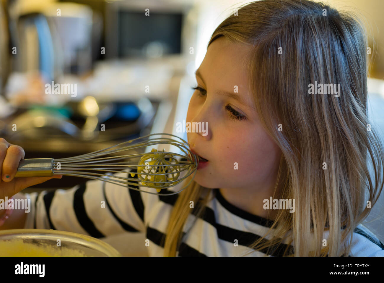 Giovane ragazza leccare cake mix da una frusta di metallo in una cucina con bokeh di fondo Foto Stock