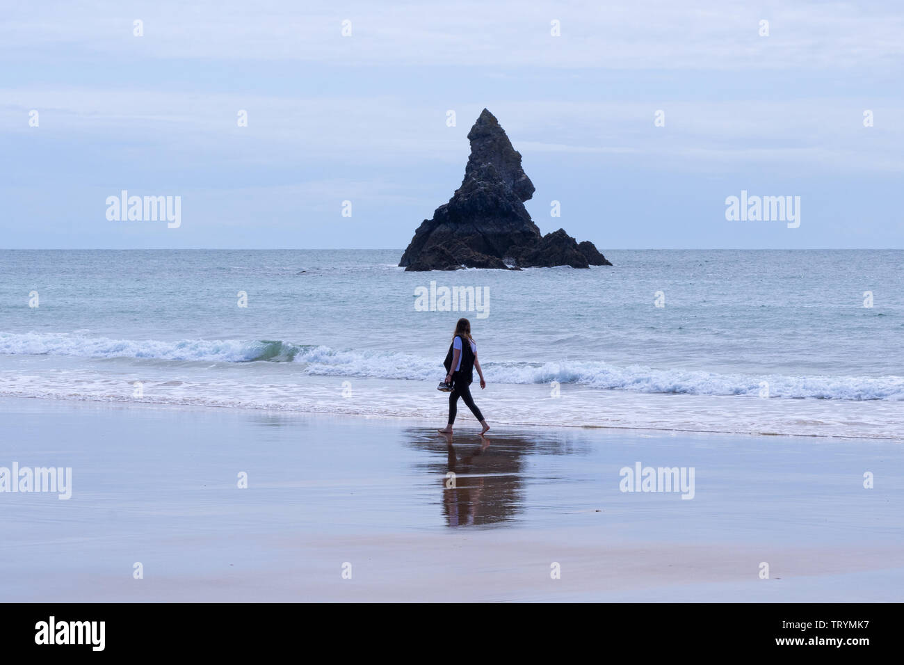 Donna di camminare sulla spiaggia in acque poco profonde che trasportano le scarpe di fronte alla chiesa Rock ad ampio Haven, Pembrokeshire, Galles Foto Stock