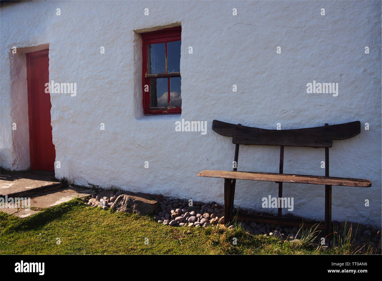 Una vista ravvicinata di Croft cottage, del rosso porta anteriore di una finestra e un esterno panchina contro il muro bianco Foto Stock