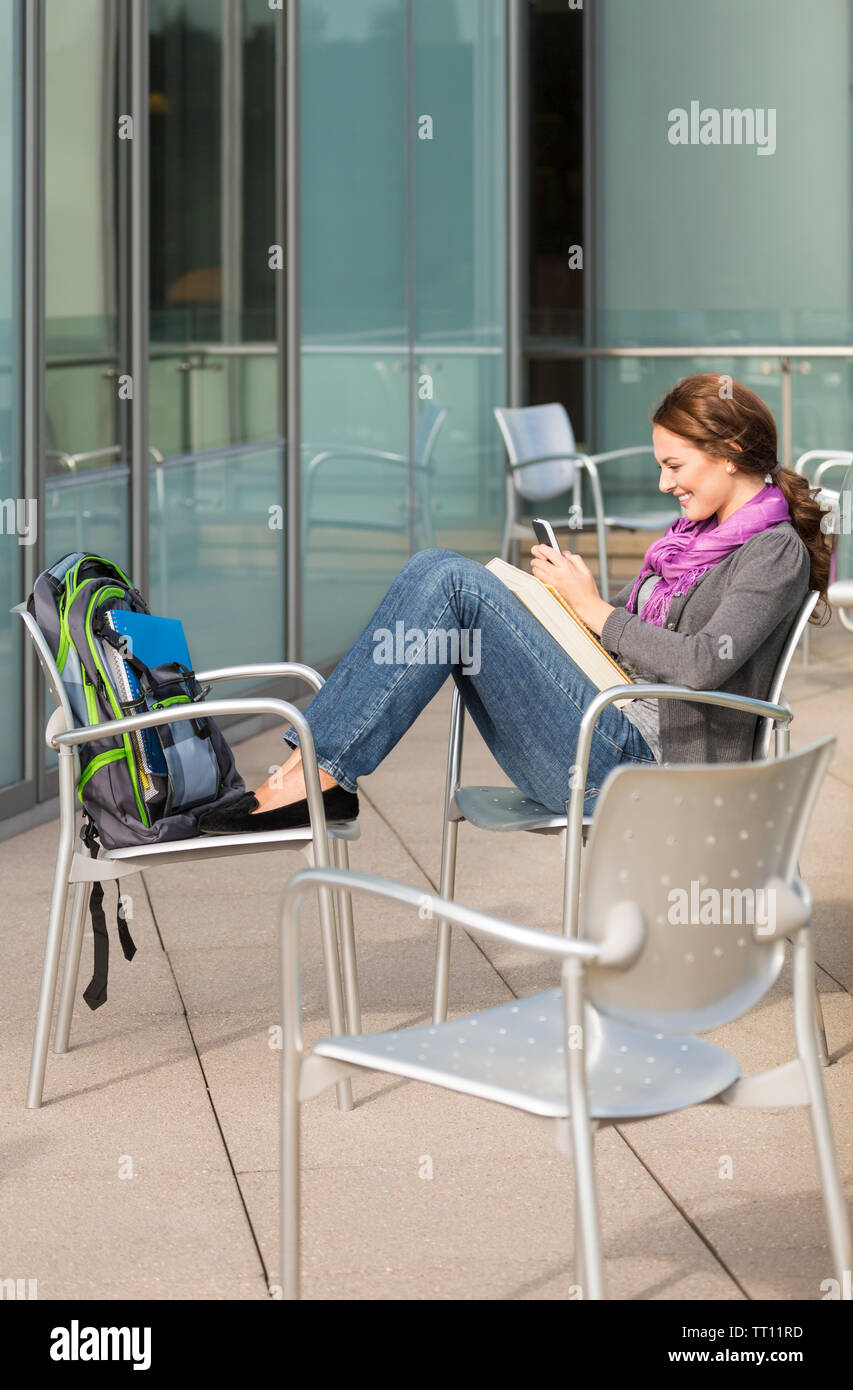 Felice, sorridente, giovane donna collegio femminile studente universitario con un telefono cellulare all'esterno del campus. Guarda come se gli SMS o utilizzando un'applicazione. Foto Stock