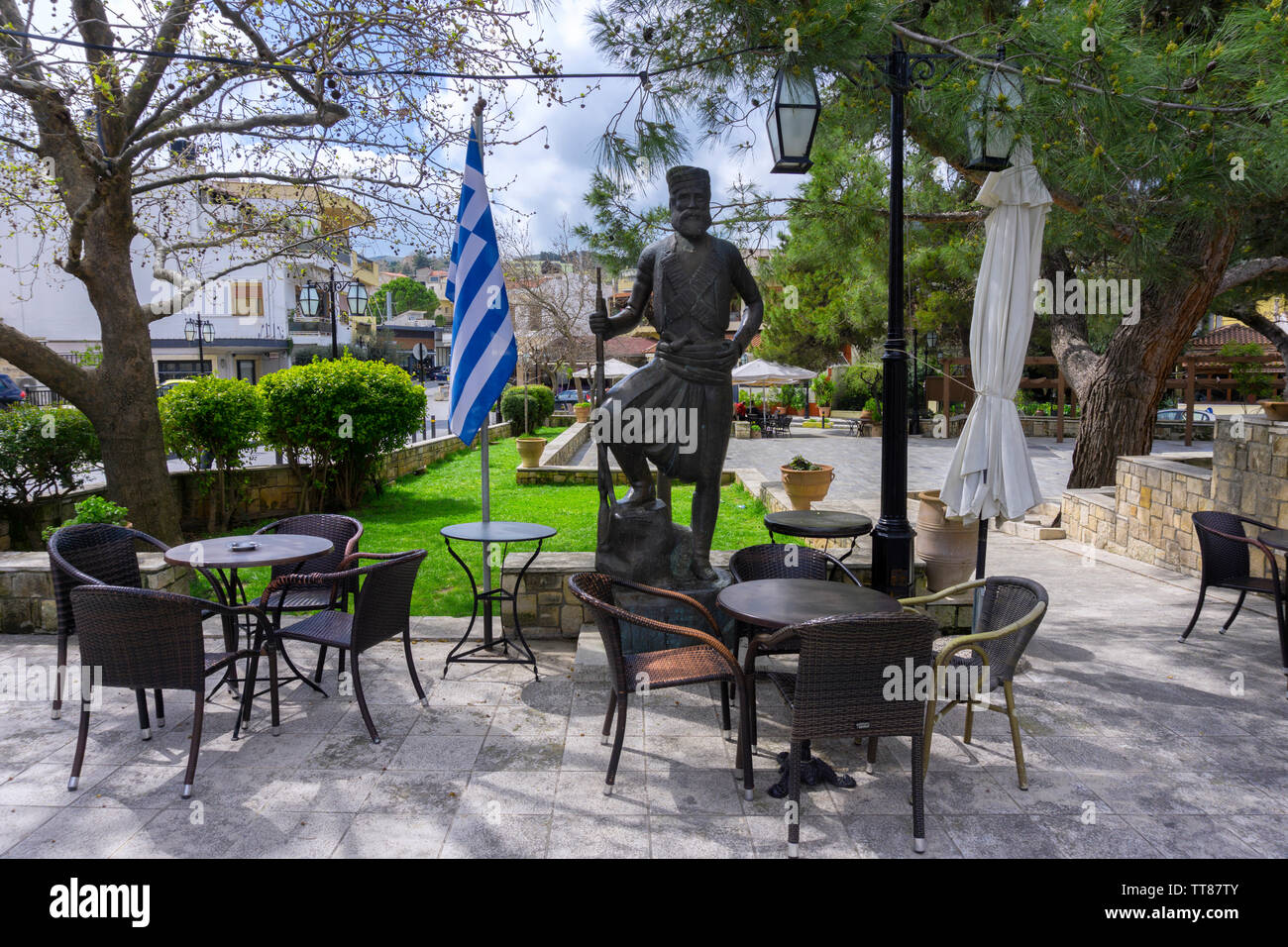 Archanes, isola di Creta / Grecia. La piazza centrale di Archanes Village con i tradizionali negozi di caffè. Statua di bronzo di un eroe cretese Foto Stock