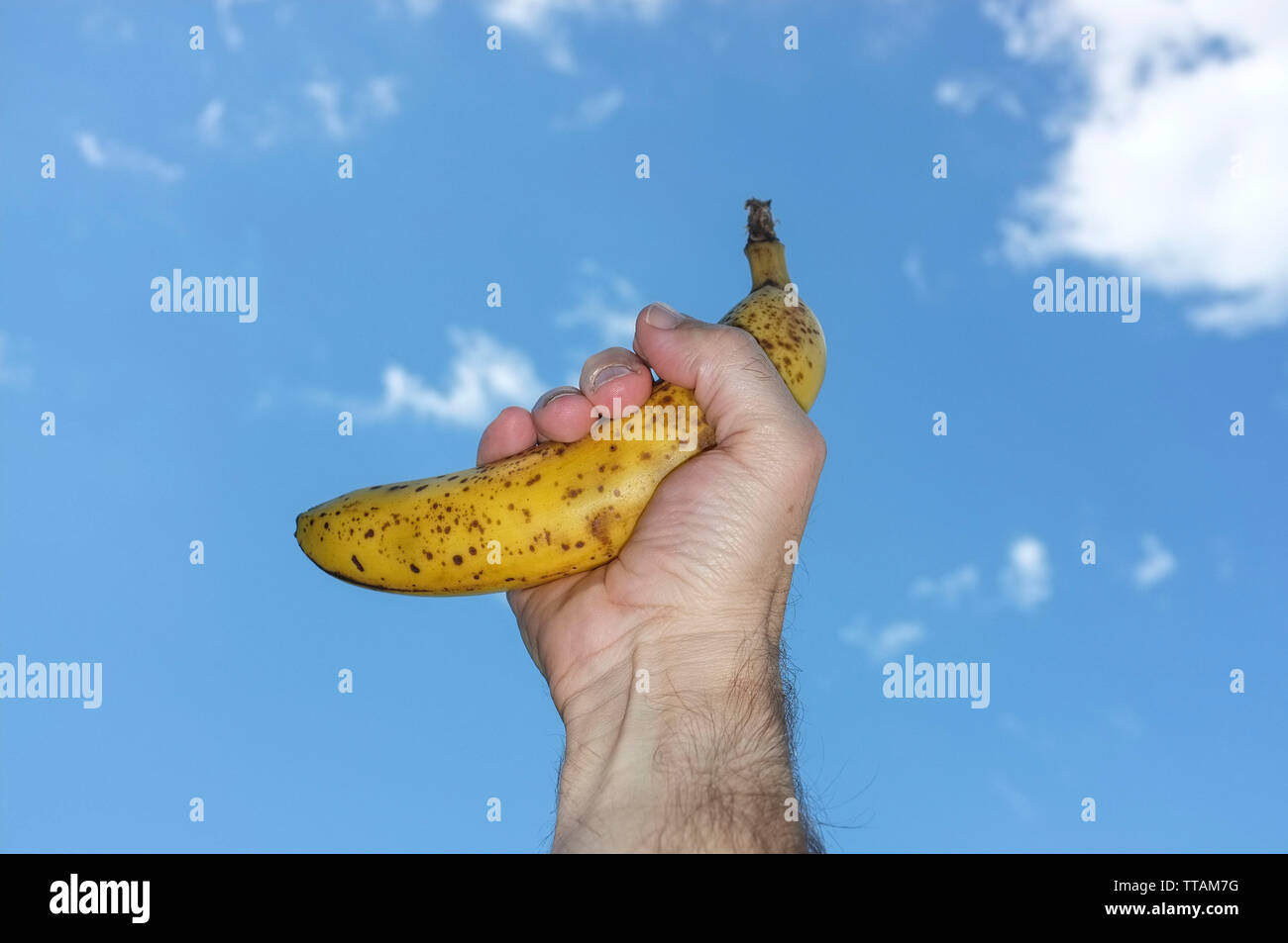 La mano di un uomo in possesso di un maturo banana giallo marrone con macchie enzimatiche chiamato Browning, contro un cielo blu con nuvole bianche. Foto Stock